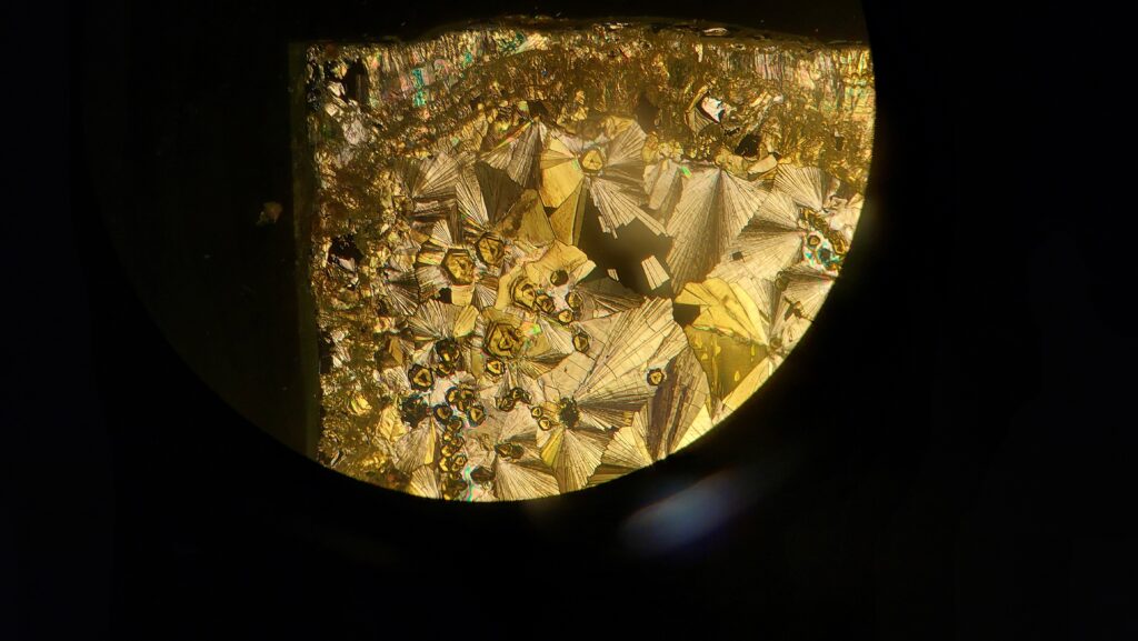 A microscopic view of a crystalline structure with a brilliant gold and metallic luster. The crystals form radiating, fan-like patterns and geometric, hexagonal shapes that overlap across the frame. Some areas show iridescent, rainbow-like highlights, all contained within a circular field of view against a dark, shadowed background.