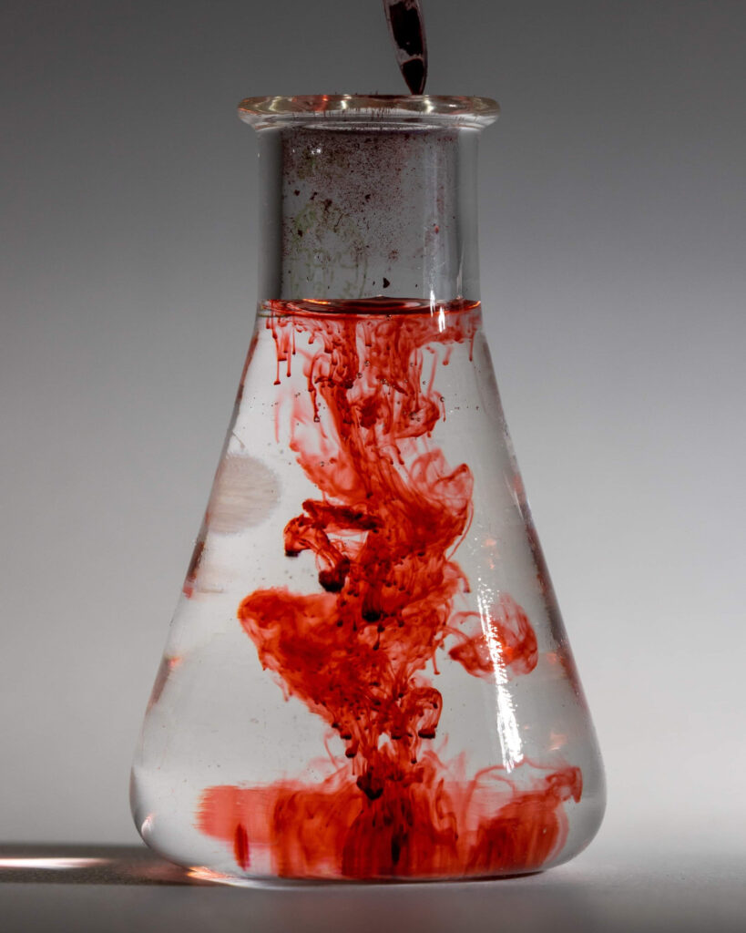 A clear glass Erlenmeyer flask filled with water sits against a neutral gray background. A dark red liquid is being dripped into the flask from above, creating blooming, cloud-like plumes of crimson ink that swirl and descend through the clear water. Small red droplets are visible near the surface, while a more concentrated mass of color settles near the bottom of the flask.