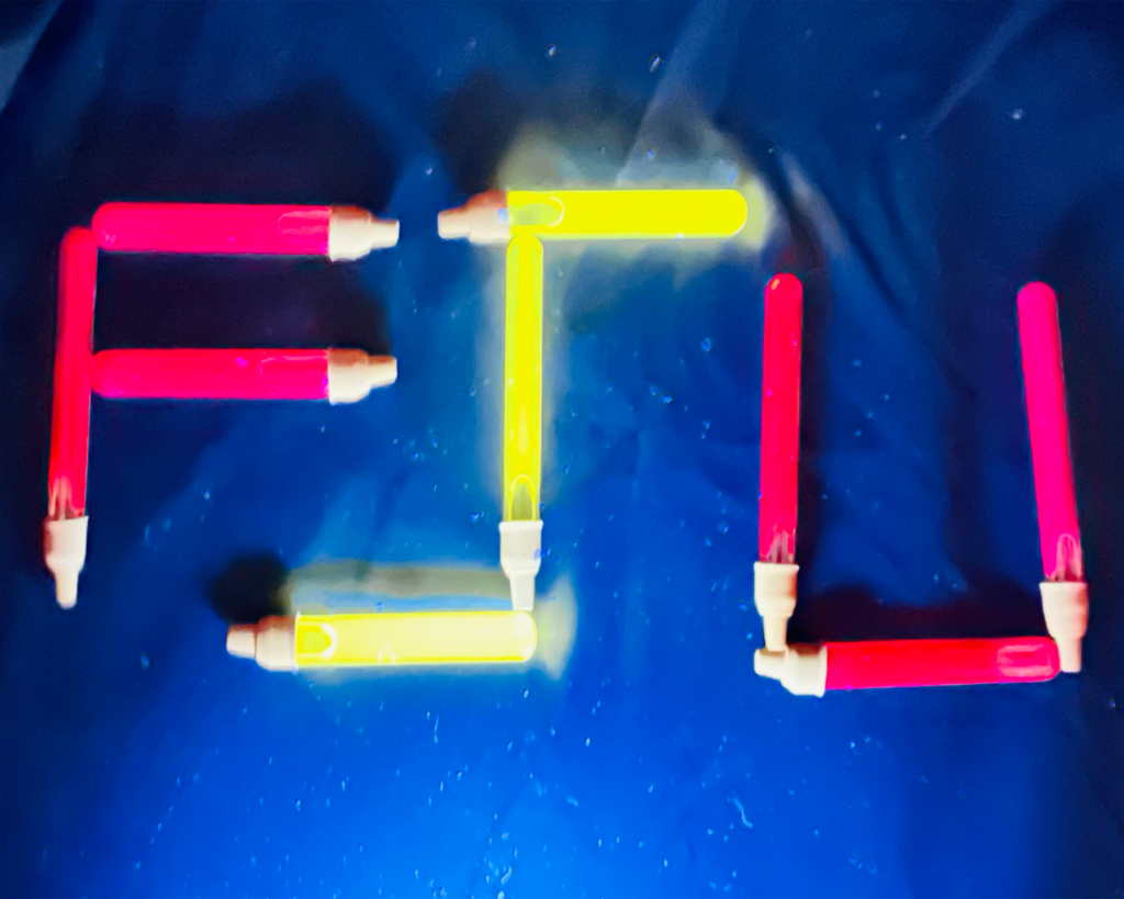 Several glowing lab vials are arranged on a blue surface to spell out the letters FSU. The letters F and U are made of vials glowing a bright, neon pink, while the S is made of vials glowing a vivid neon yellow. The blue background is slightly textured and reflective, catching the glow from the colored liquids.