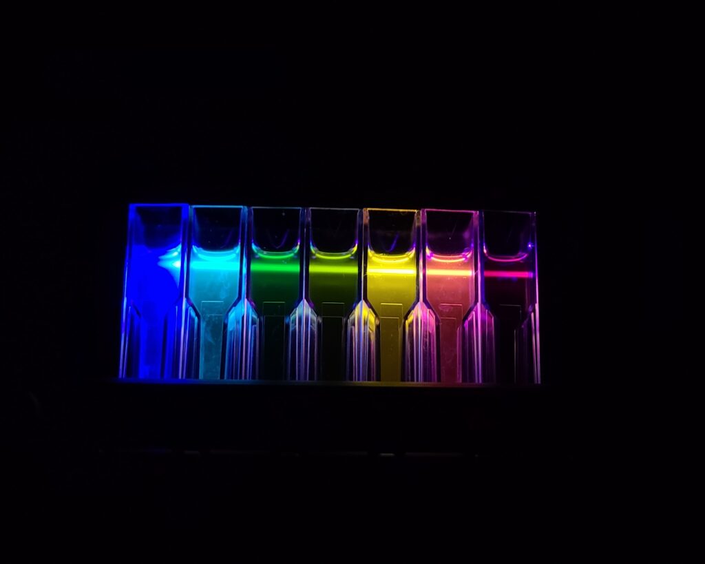 A row of seven clear laboratory cuvettes filled with glowing liquids, arranged to form a rainbow spectrum against a pitch-black background. From left to right, the liquids transition through vibrant shades of royal blue, cyan, emerald green, lime green, bright yellow, orange-red, and deep magenta. Each container is illuminated from within, casting a sharp, colorful glow that makes the chemicals look like a neon light display.