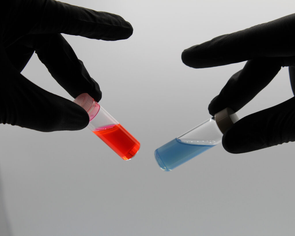 Two hands in black nitrile gloves are holding up small glass vials against a plain white background. The vial on the left contains a bright reddish-orange liquid, while the vial on the right contains a light blue liquid. Both vials are tilted toward each other in the center of the frame, showing the colored solutions inside.