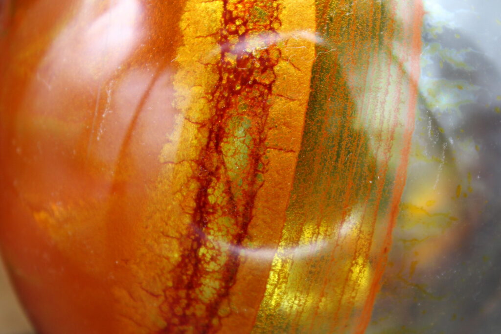 A extreme close-up of a smooth, rounded surface showing vertical bands of color. The middle of the image features a bright gold stripe with a dark, reddish-brown cracked pattern running down it. To the left, the surface is a solid orange, and to the right, it is a semi-transparent grey with thin orange lines. A blurry, circular reflection of light is visible in the center.