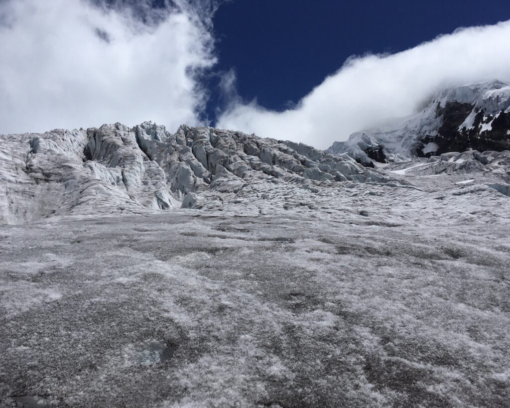 A wide-angle photograph of a vast, jagged glacier under a deep blue sky. The foreground and middle ground are filled with uneven, grey-white ice and compacted snow, showing a rough and weathered texture. In the background, the glacier rises into steep, fractured ice cliffs and snow-covered mountain peaks, partially shrouded by wispy white clouds. The scale of the ice formation conveys a sense of cold, silent isolation in a high-altitude environment.