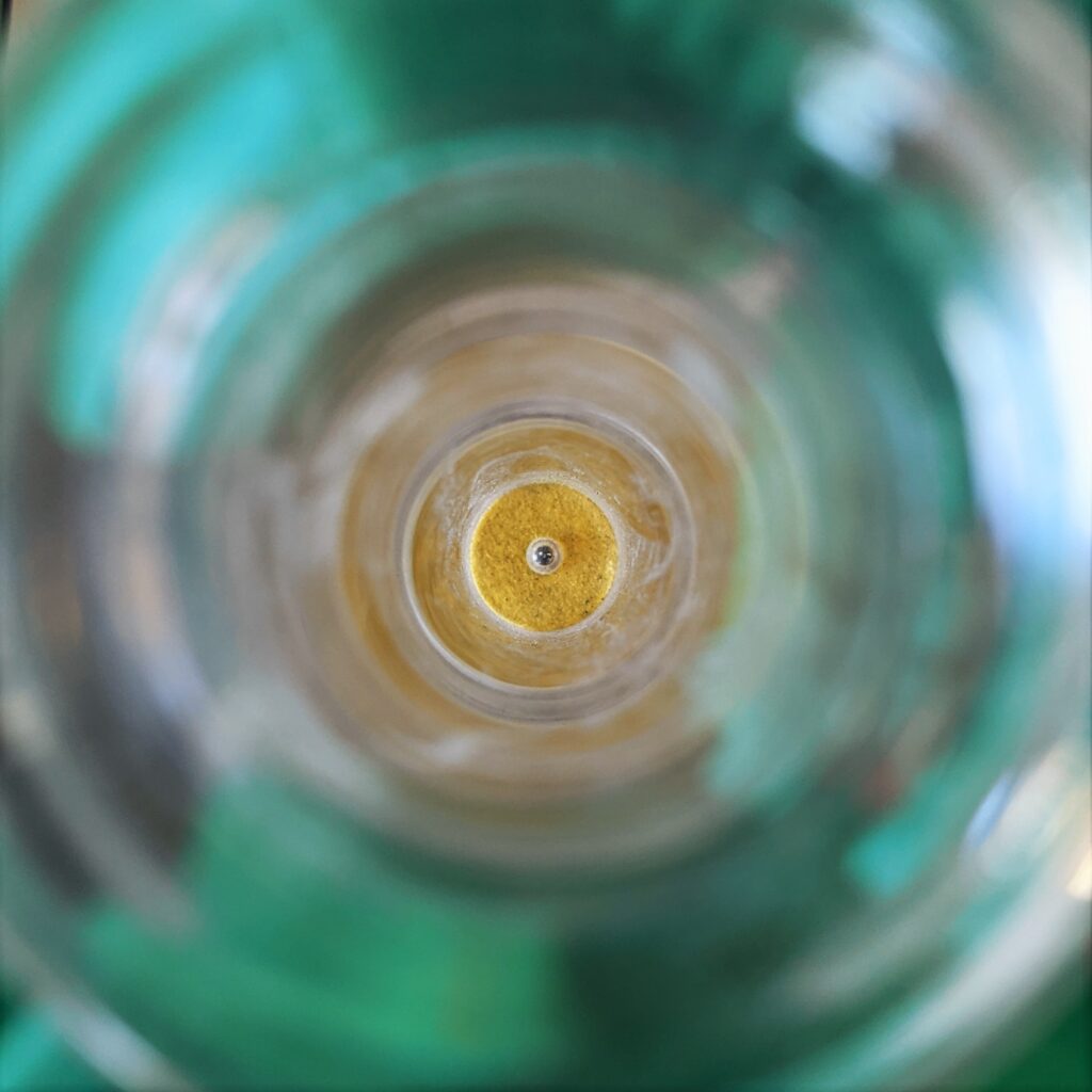 A top-down photograph looking into a circular glass apparatus. At the center, a tiny, dark metallic sphere sits atop a vibrant yellow, textured circular base. Surrounding this focal point are concentric rings of blurry glass and liquid, with a soft, out-of-focus teal or turquoise background visible through the transparency of the container. The lighting is centered, creating a tunnel-like effect that draws the eye directly to the small sphere in the middle.