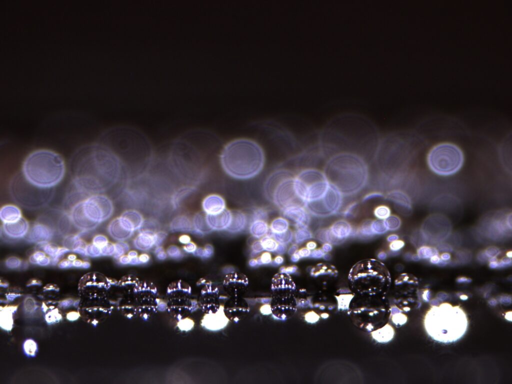A macro photograph showing an OER (Oxygen Evolution Reaction) electrocatalyst in action. Small, highly reflective oxygen bubbles are being produced on the surface of a glassy carbon electrode modified with cobalt nanocarbide. The bubbles in the foreground are in sharp focus, appearing as perfect silver spheres that mirror the surrounding light. In the background, dozens of out-of-focus oxygen bubbles create a soft, ethereal bokeh effect, appearing as glowing lavender and white rings against a deep, dark liquid environment.