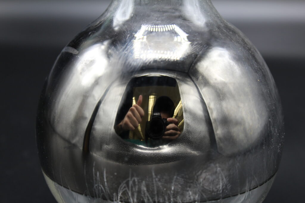 A photograph of a round, silver-colored flask with a highly reflective, metallic surface. In the center of the flask, a clear reflection is visible, showing a person holding a camera and giving a thumbs-up. The metallic coating appears slightly distressed with fine vertical scratches and faint white crystalline patterns at the base. Overhead laboratory lights are reflected as bright, hexagonal shapes on the curved upper surface of the vessel.