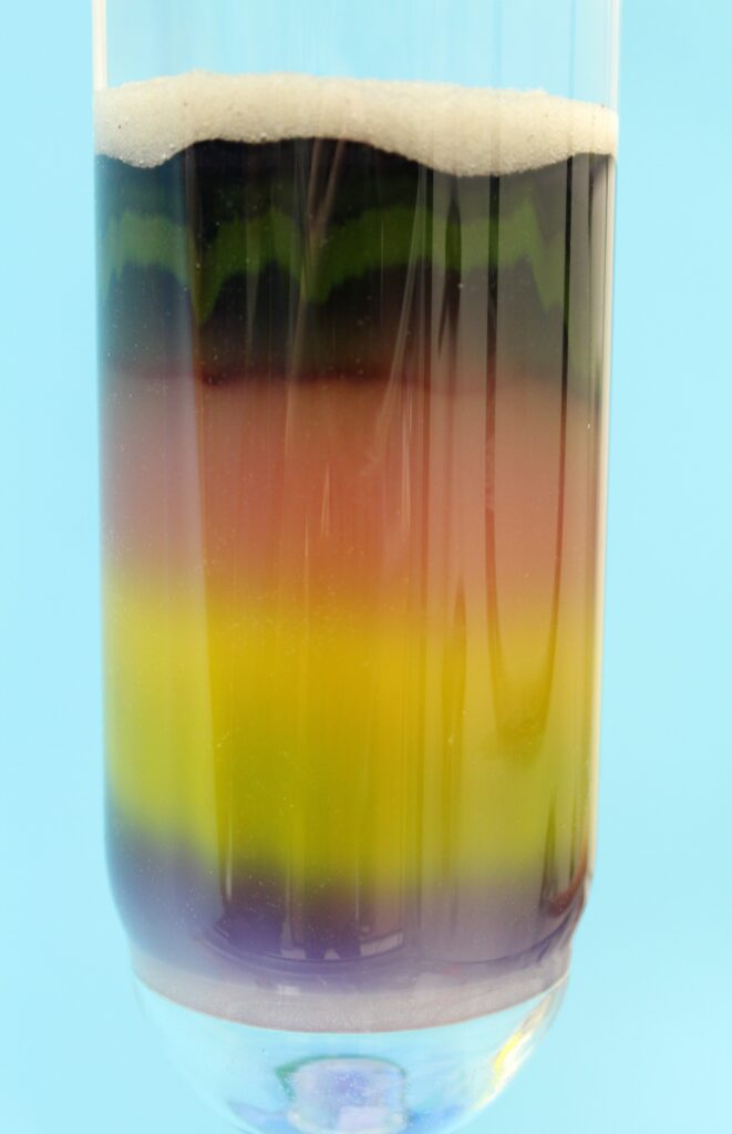 A photograph of a tall glass cylinder containing a colorful density gradient. Layers of liquid are stacked from bottom to top in shades of purple, yellow-green, bright yellow, orange-red, and a dark, nearly black top layer. A thick, white foam or granular substance sits at the very top of the stack. The glass is set against a solid light blue background.