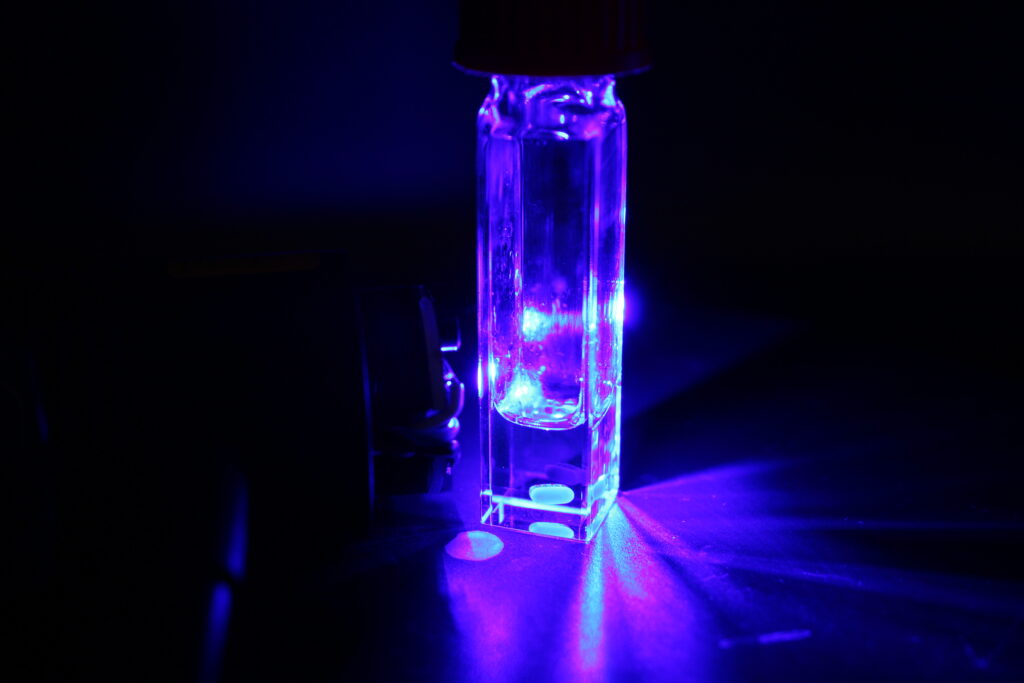 A photograph of a tall, slender glass cuvette containing a clear liquid, brilliantly illuminated by a ultraviolet or blue light source. The cuvette glows with an intense, electric violet-blue light, casting long, dramatic shadows and radial light beams across the dark surface it sits on. Two bright "hotspots" of fluorescence are visible near the bottom of the vessel, while the background remains in deep shadow, emphasizing the luminosity of the sample.