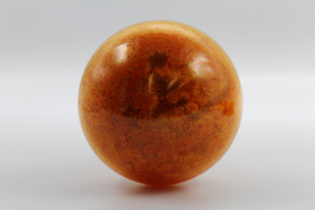 A photograph of a smooth, polished sphere sitting on a clean white surface. The sphere features a rich, amber-orange color with a speckled, granular internal texture and subtle red highlights. A soft reflection of light glints off its glossy top-left surface, while the overall lighting creates a gentle shadow at its base against the plain, light gray background.