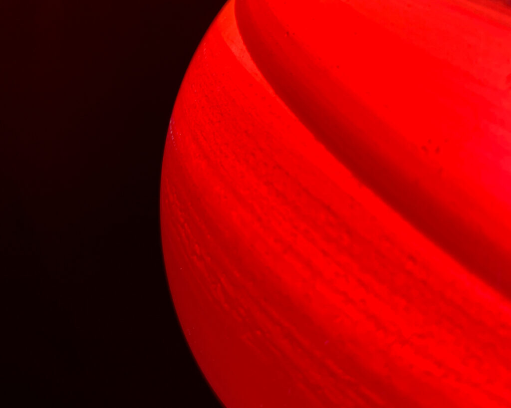 A close-up photograph of a bright red, curved object set against a solid black background. The object shows subtle horizontal textures and a smooth, rounded surface with a soft, bright highlight along the upper right edge. Its shape and glossy finish suggest a large spherical or cylindrical form, though only a portion of its side is visible.