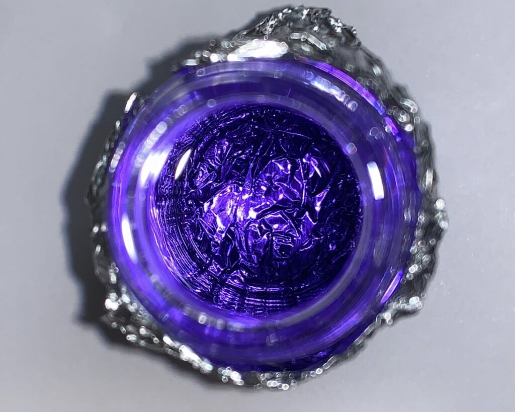 A top-down photograph of a small glass jar or container resting on a crinkled piece of aluminum foil. The jar is filled with a deep, vibrant violet liquid that appears to be fluorescing under a light source. The reflections on the liquid's surface and the glass rim create bright highlights, while the crumpled foil underneath adds a metallic, shimmering texture around the circular base of the jar.