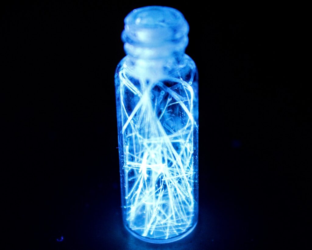 A small glass vial filled with needle-like crystalline structures that glow with an intense, electric blue bioluminescence or fluorescence. The glowing fibers are tangled and crisscrossed inside the container, casting a bright blue light that illuminates the glass against a pitch-black background. The light is most concentrated at the center of the vial, where the crystals are most dense.