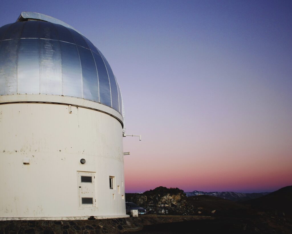 A photograph of a white, cylindrical observatory building topped with a large, metallic silver dome. The structure sits on a dark, rocky foundation against a twilight sky that gradients from deep blue at the top to a soft pink and orange at the horizon. In the distant background, the dark silhouettes of mountain ranges are visible under the fading light.