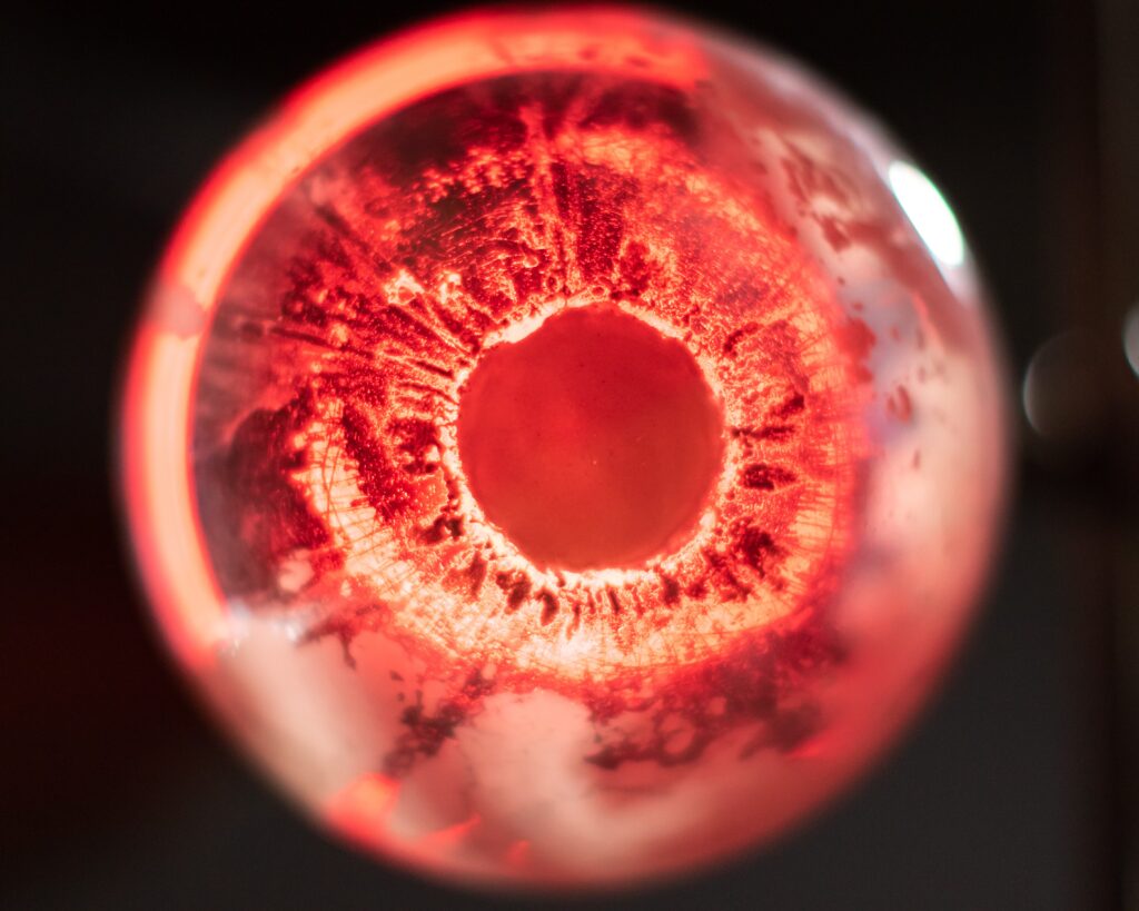 A close-up, top-down photograph of a glowing red sphere with a dark circular center. The interior surface is marked by dense, radiating lines and dark, irregular patches that create a texture similar to an eye's iris or a biological cell. The light source is positioned behind the object, creating a bright crimson glow that fades into a soft, out-of-focus blur around the edges.
