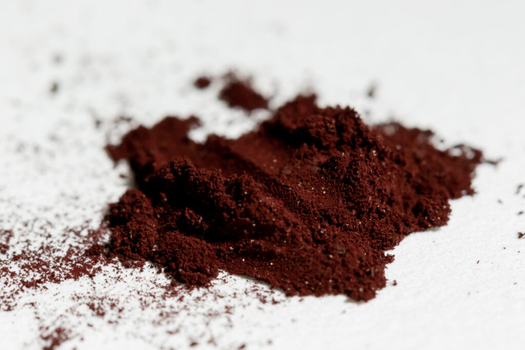 A photograph of a small pile of fine, deep maroon or burgundy powder resting on a white, textured surface. The powder has a rich, earthy pigment and a clumpy, granular consistency, with tiny stray particles scattered across the surrounding white background. The focus is sharp on the center of the pile, highlighting its matte, velvety texture.