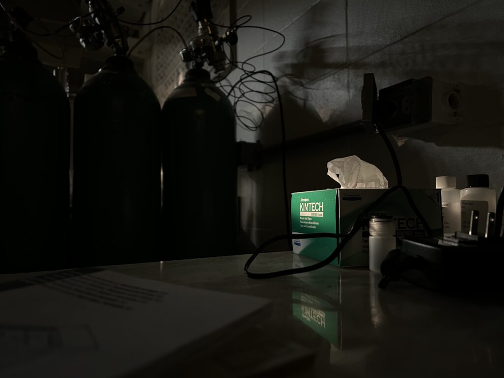 A dimly lit laboratory space with three large black gas cylinders standing in the shadows to the left. A single spotlight highlights a green box of Kimtech science wipes on a reflective white countertop. A tangled black power cord and several small white reagent bottles are visible nearby against a grey cinderblock wall.