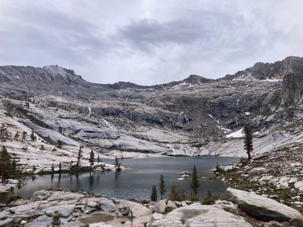 A high-altitude alpine lake surrounded by steep, grey rocky mountains. Several waterfalls flow down the smooth rock faces into the lake, and sparse pine trees grow along the rocky shore under an overcast grey sky.