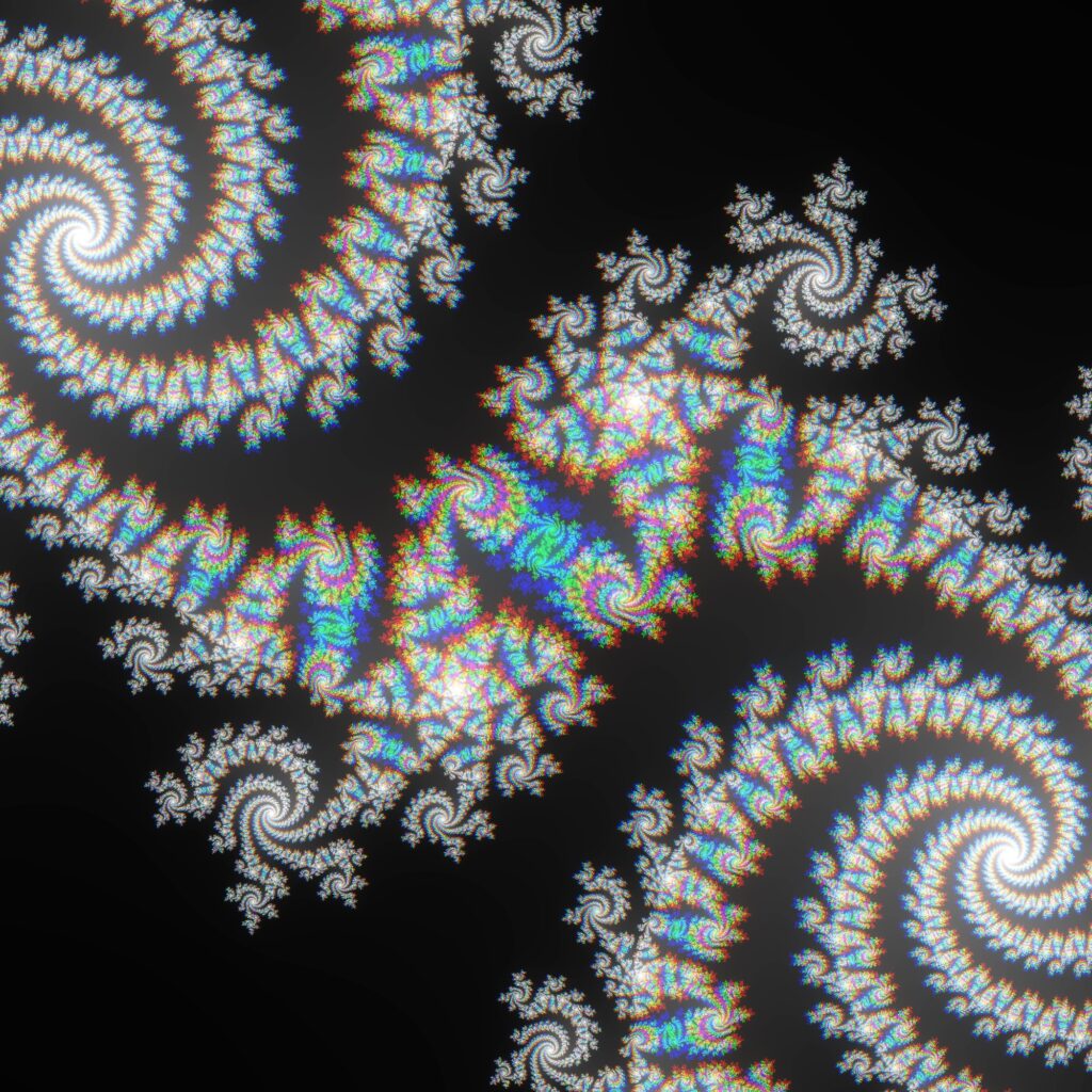 A vibrant, multicolored fractal design featuring intricate, swirling patterns that resemble spiraling galaxies or nautilus shells. The patterns consist of bright rainbow hues with cyan, magenta, yellow, and green, set against a stark black-to-grey gradient background.