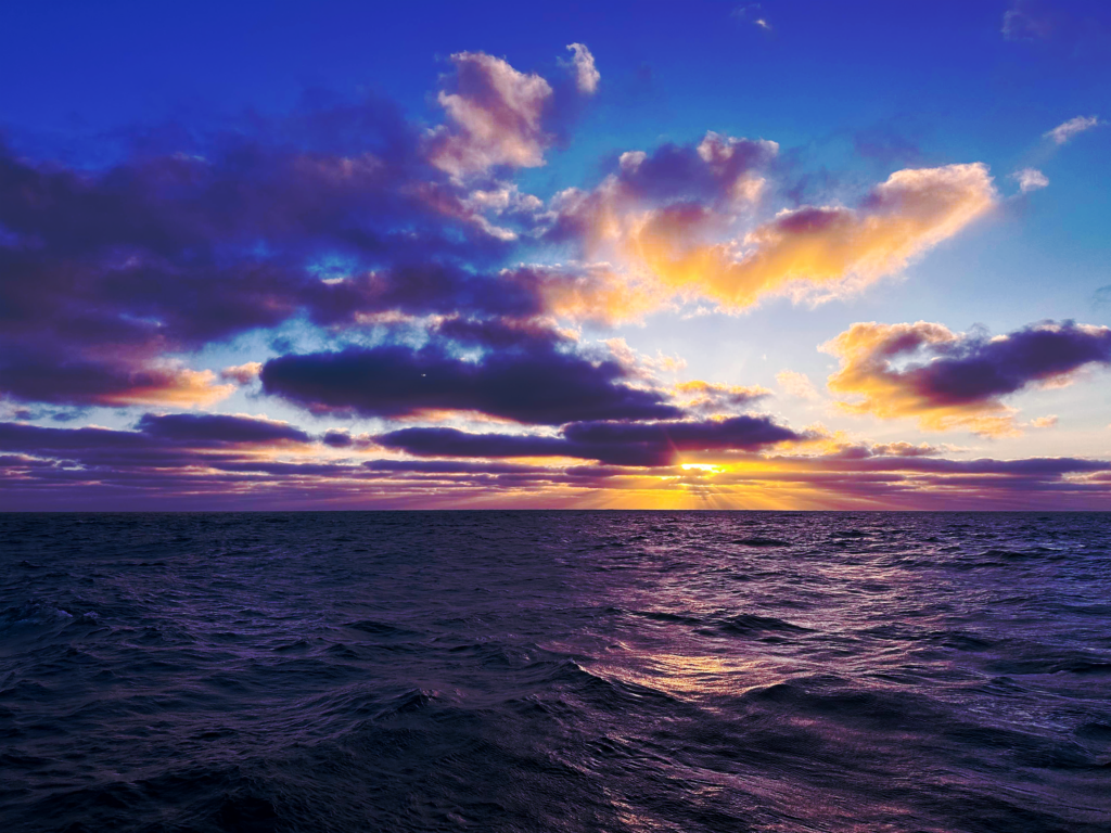 A landscape photograph of a sunset over the ocean. The sun sits low on the horizon, casting yellow and orange rays through a layer of dark, horizontal clouds. Above, large scattered clouds are tinged with pink and gold against a deep blue sky. The foreground shows the textured surface of dark purple and blue waves reflecting the light from the horizon.